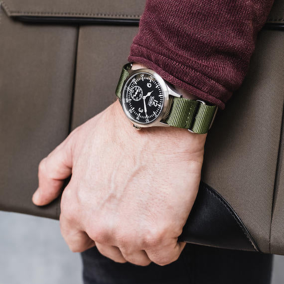 Hand wearing a Techné Merlin watch with a green strap on a brown leather surface