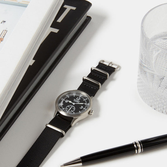Techné Merlin wristwatch with silver accents on a white surface with a pen and notebook.